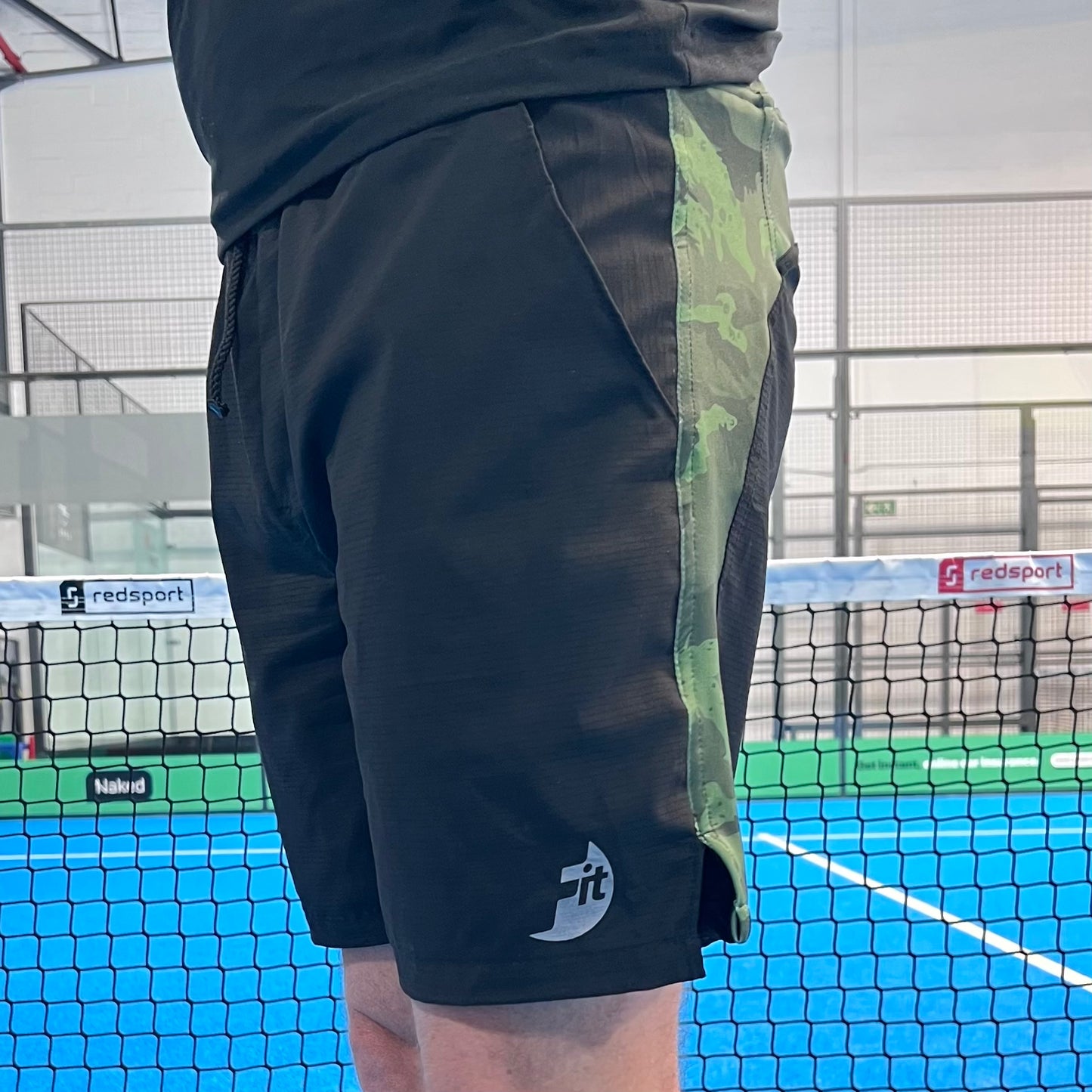 Black athletic shorts with green camouflage side panel on a padel court